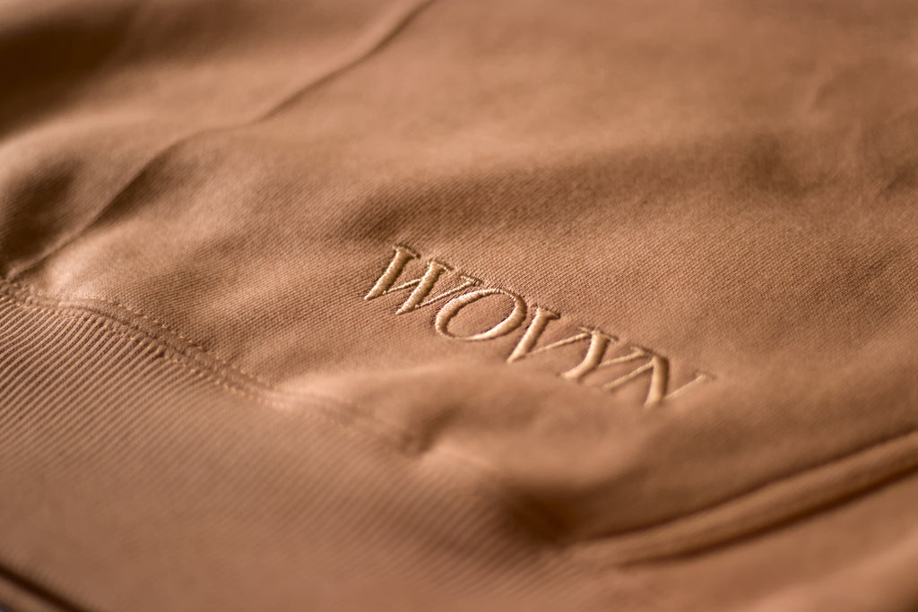 Macro detail of tonal WOVYN logo embroidery on the Soft Sand hoodie, showing heavyweight fleece texture